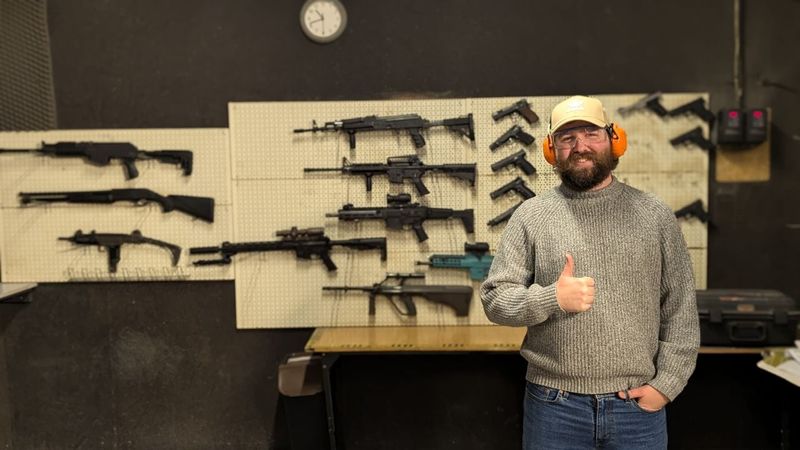 A thumbs up in ear defenders and safety glasses in front of a wall of rifles at a gun range
