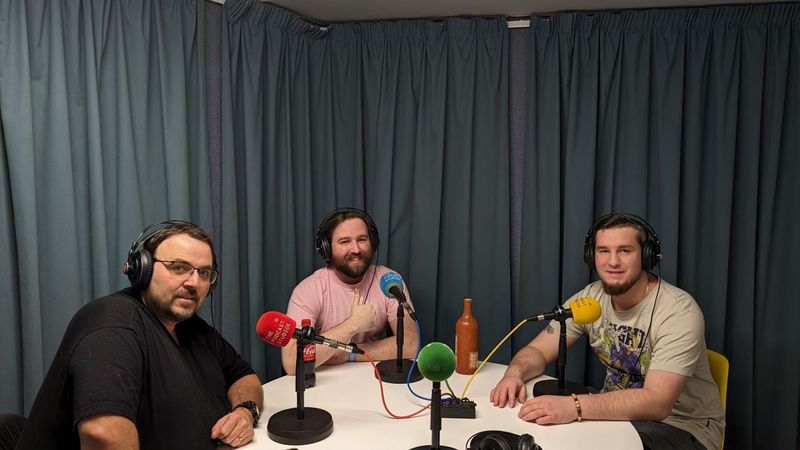 Three of us in headphones around a white table of colourful microphones in a podcast studio