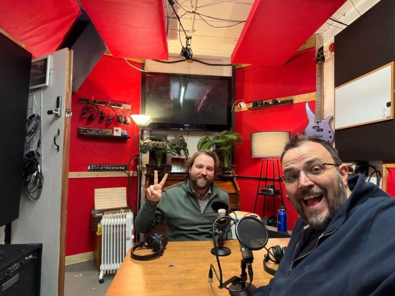 A selfie in a red-walled recording studio, with a peace sign from across the table