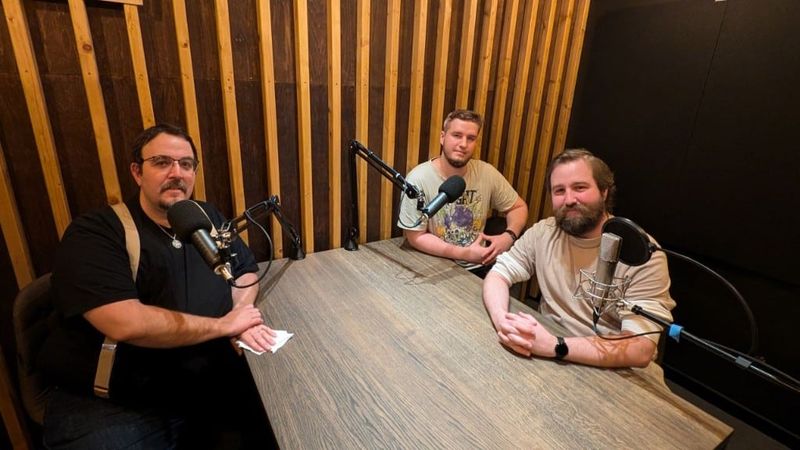 Three of us at a wooden table with microphones in a studio with slatted wooden walls