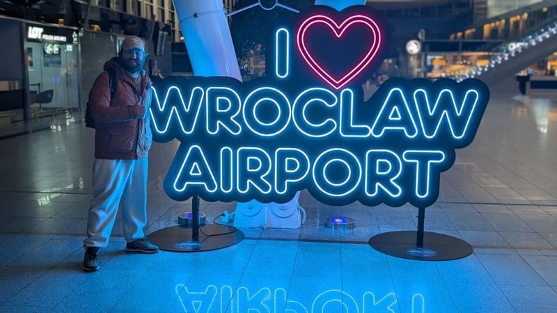 Giving a thumbs up beside a glowing neon "I love Wrocław Airport" sign